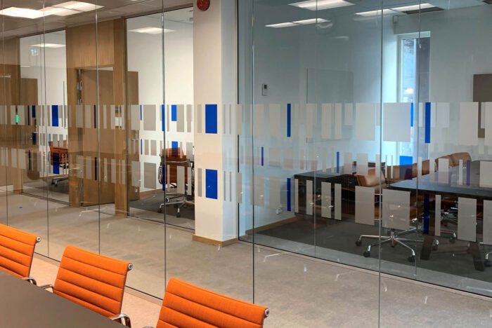En glassvegg i et kontorlandskap har en horisontal stripe med dekorativ folie. Dekoren består av et abstrakt mønster med frostede og blå loddrette rektangler.